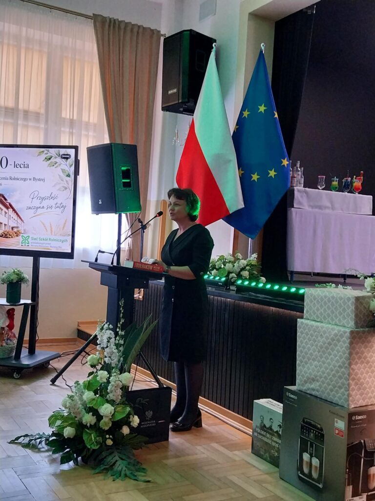 dr hab. inż. Barbara Krochmal-Marczak, prof. PANS, kierownik Zakładu Produkcji i Bezpieczeństwa Żywności stoi przy mównicy z tyłu flaga Polski i UE, scena, prezenty, stół na pokaz drinków