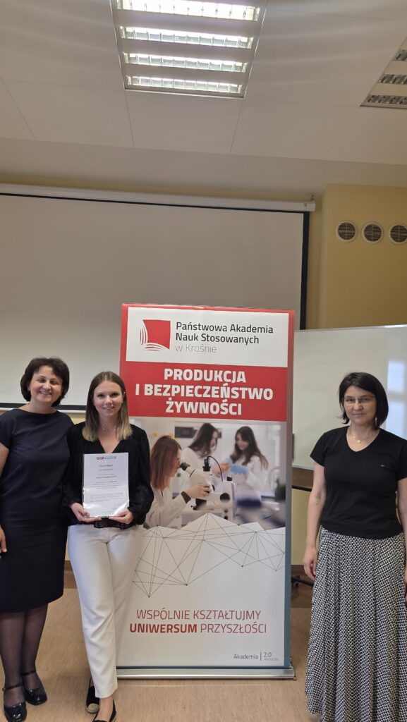 Studentka z certyfikatem stoi obok baneru kierunku Produkcja i bezpieczeństwo żywności po prawej stoi dr Dykiel, po lewej prof. Krochmal-Marczak