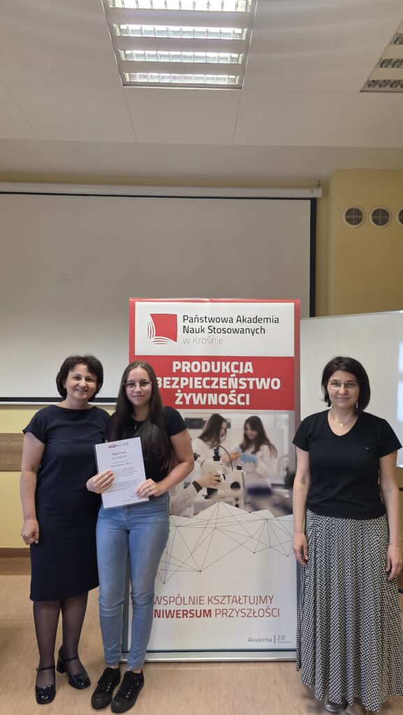 Studentka z certyfikatem stoi obok baneru kierunku Produkcja i bezpieczeństwo żywności po prawej stoi dr Dykiel, po lewej prof. Krochmal-Marczak