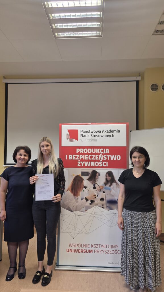 Studentka z certyfikatem stoi obok baneru kierunku Produkcja i bezpieczeństwo żywności po prawej stoi dr Dykiel, po lewej prof. Krochmal-Marczak
