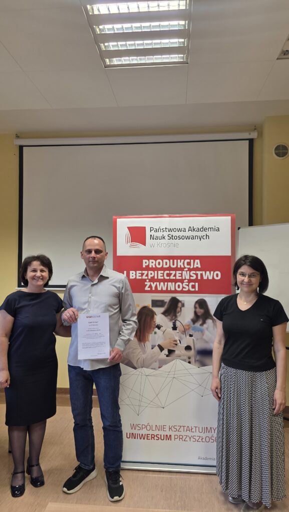 Student z certyfikatem stoi obok baneru kierunku Produkcja i bezpieczeństwo żywności po prawej stoi dr Dykiel, po lewej prof. Krochmal-Marczak