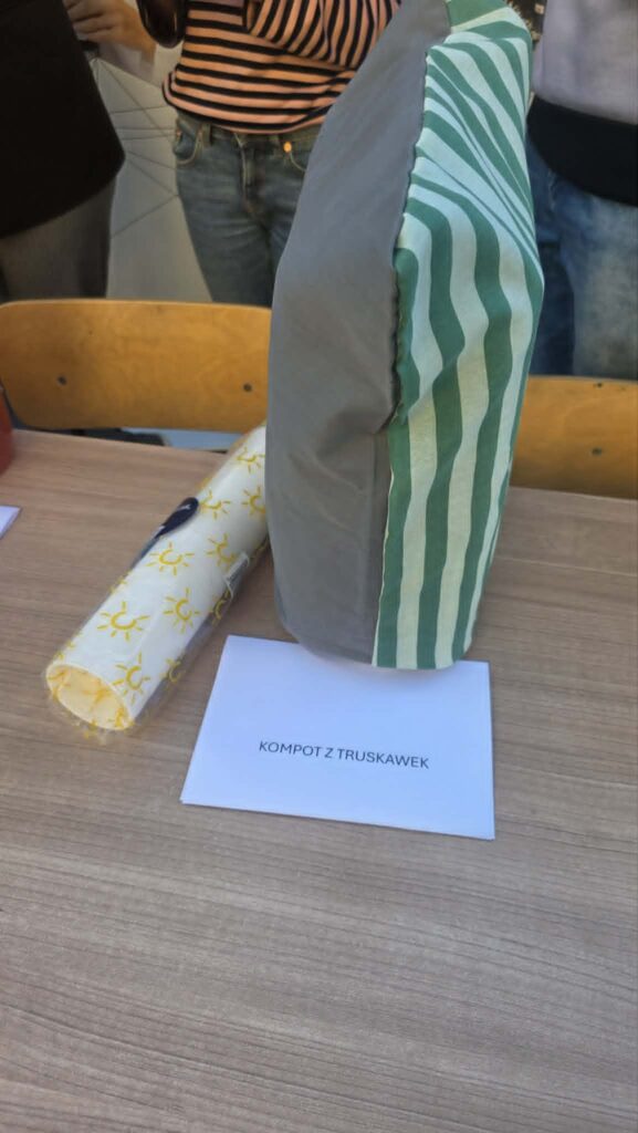 Na stole w słoju z korkiem przykryty pokrowcem w pasy stoi napój z napisem kompot z truskawek