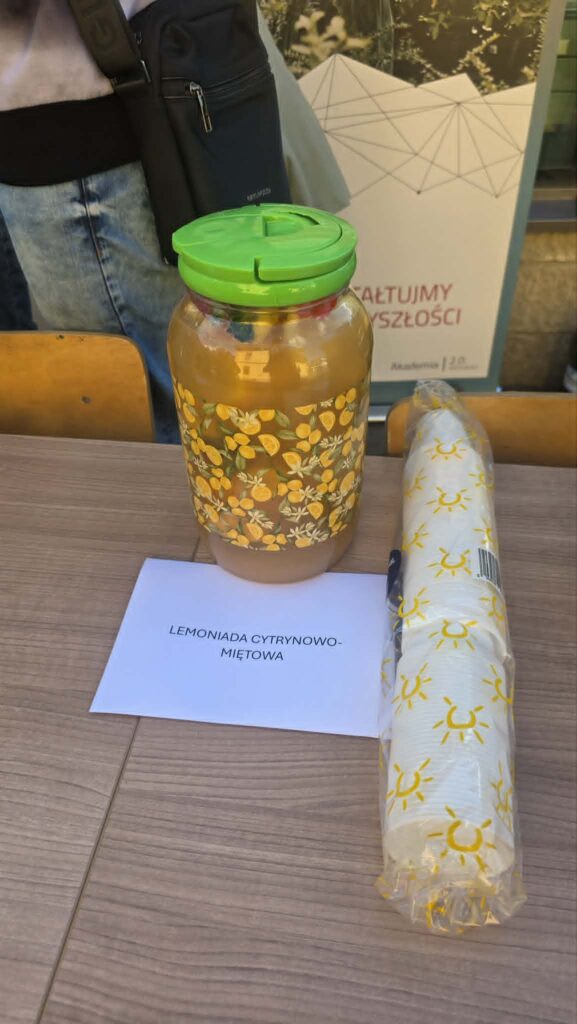 Na stole w słoju z korkiem stoi napój z pomarańczami w środku, napis lemoniada cytrynowo miętowa