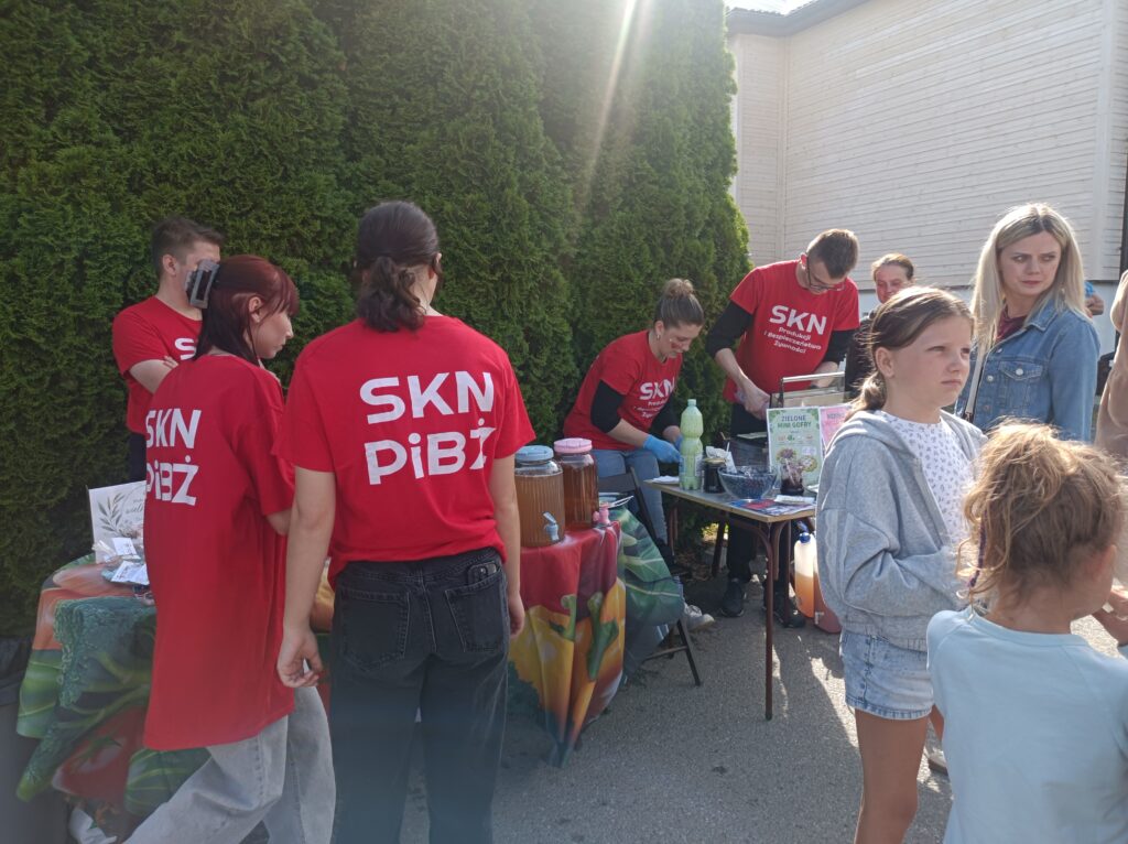 Stoisko studentów, studenci w czerwonych koszulkach z napisem SKN PIBŻ stoją przodem i tyłem, przygotowują produkty. Z boku po prawej ludzie-uczestnicy