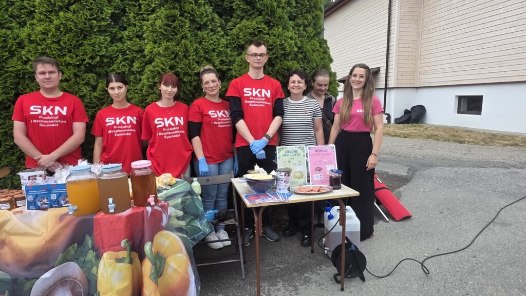 Na tle stoiska z produktami, napojami stoją studenci biorący udział w wydarzeniu.