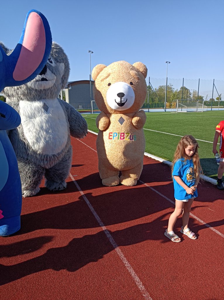 Na bieżni stadionu idą dwie duże maskotki, Koala i miś
