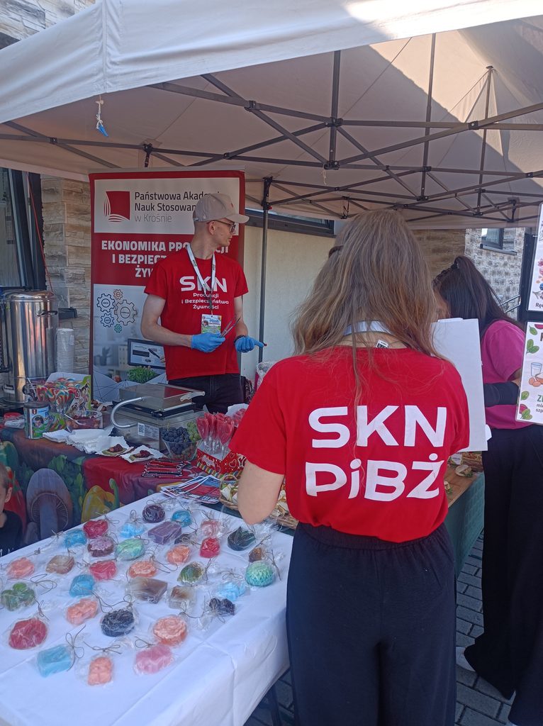 Przy stoisku koła SKN PIBŻ stoi studentka odwrócona plecami w czerwonej koszulce z napisem SKN PIBŻ, z tyłu student i banner wszystko dzieje się przy stole promocyjnym