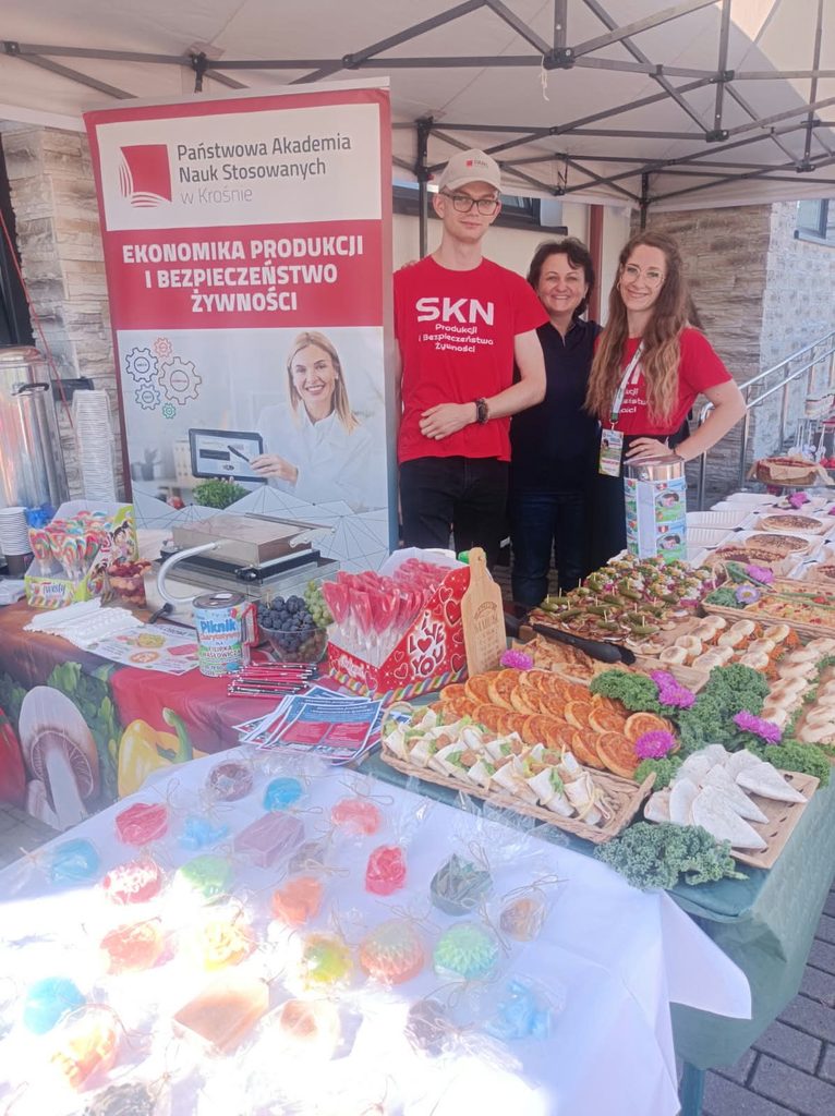 na pierwszym planie na stole leża kolorowe mydełka, lizaki, owoce gofrowania, od lewej baner kierunku EPIBŻ i 3 osoby