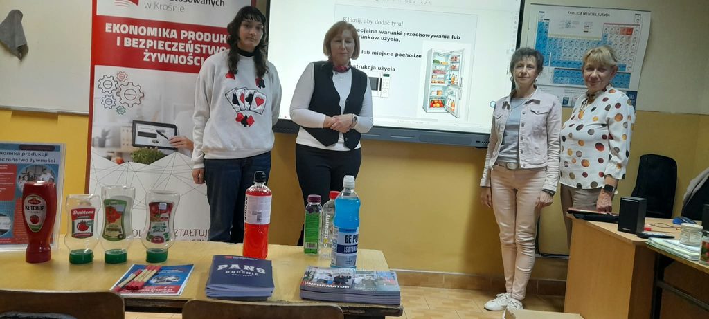 Nauczyciele ze szkoły-po prawej stoją, po drugiej stronie prelegentka i uczennica. za nimi baner kierunku EPIBŻ i tablica aktywna z prezentacja wykłądu. na pierwszym planie ławka a na niej opakowania po produktach spożywczych, informatory, zeszyty i ołówki