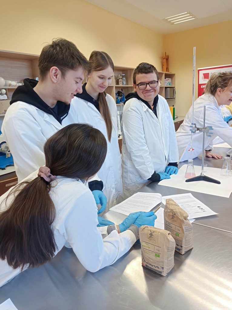 uczniowie przy stole laboratoryjnym badają mąkę i jej kwasowość, na stole stoi mąka w torebkach, biureta na stojaku, kolba z próbką