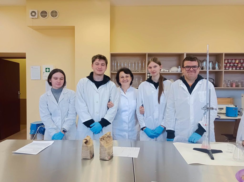 Grupa uczniów z prowadzącą prof. na tle półek ze szkłem karafki, zaparzaczce i eksponatami w laboratorium np. jaja ptaków, próbki nasion