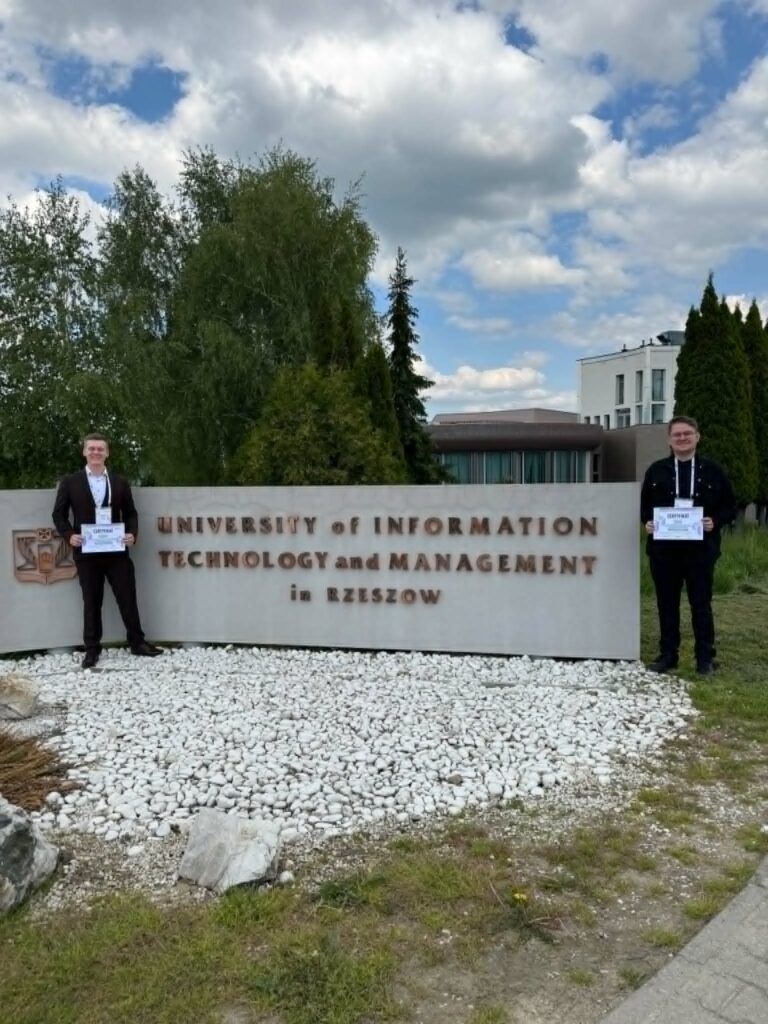 dwóch studentów stoi koło szarego murka z nazwą uczeni gościnnej