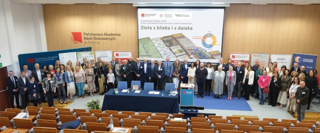 Zdjęcie na auli wszystkich uczestników konferencji. W tle banery i ekran z tytułowym slajdem konferencji.