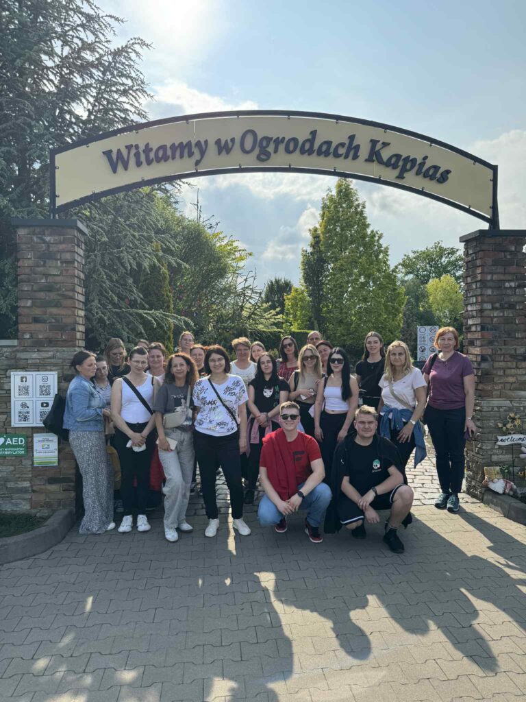 Cała grupa studentów przy bramie wejściowej do ogrodów z napisem Witamy w Ogrodach Kapias