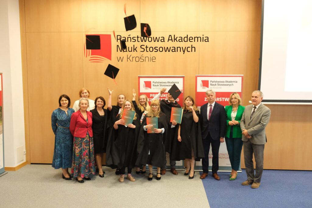 Wszyscy absolwenci z władzami i nauczycielami na tle baneru kierunku i logo uczelni rzucają birety do góry