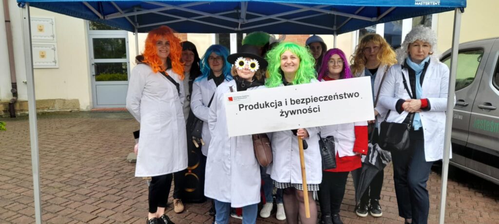Studenci kierunku PIBŻ w kolorowych perukach i napisem kierunku stoją pod namiotem