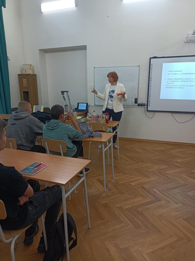Prowadząca warsztaty trzyma rekwizyty produktów, tłumaczy, uczniowie w ławkach prezentacja na tablicy