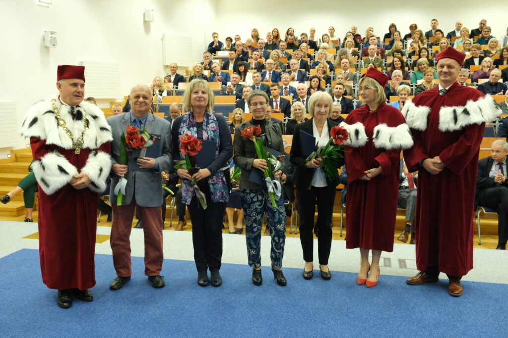 Wyróżnieni nauczyciele wraz z władzami Uczelni w tle aula z gośćmi i nauczycielami