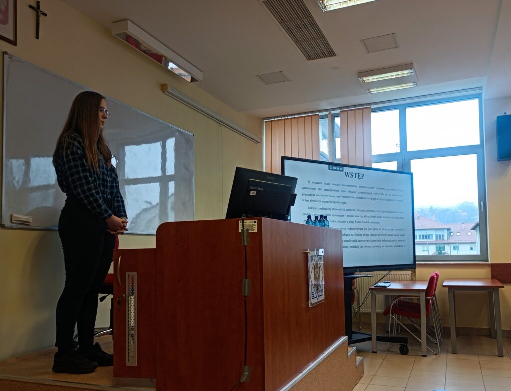 Studentka PIBŻ referuje swoje wystąpienie na podeście przy komputerze, za biurkiem