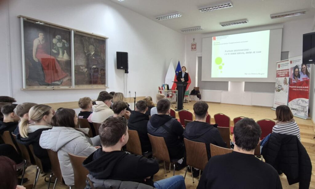 Scena, slajd z wykładem po prawej banery uczelni i kierunku, prowadząca głosi wykład na pierwszym planie uczniowie siedzą na krzesełkach tyłem