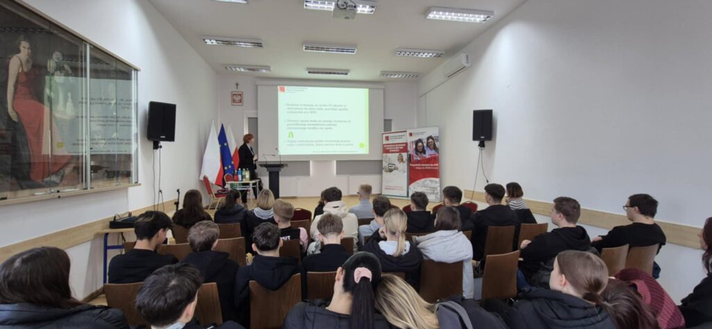 Scena, slajd z wykładem po prawej banery uczelni i kierunku, prowadząca głosi wykład na pierwszym planie uczniowie siedzą na krzesełkach tyłem