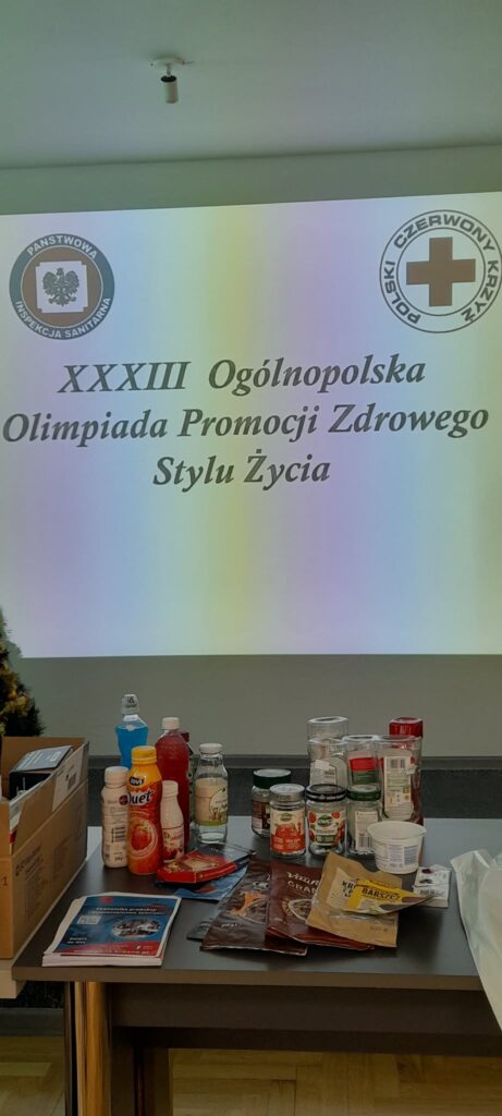 Tytuł olimpiady z rzutnika, z przodu stół z przykładowymi etykietami i produktami