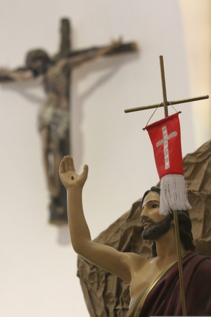 zdjęcie Jezusa który trzyma krzyż i czerwoną chorągiew znak zmartwychwstania. W tle ukrzyżowany Jezus na sianie. 