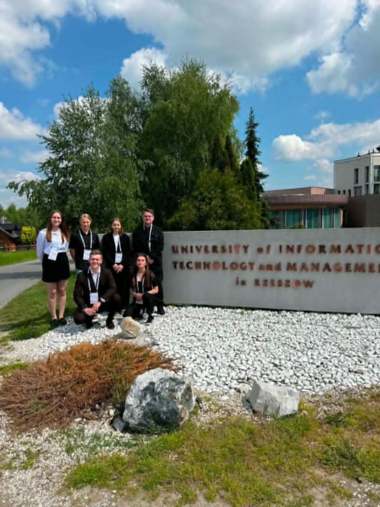 grupa studentów stoi przy dużym murze z nazwa uczelni gościnnej