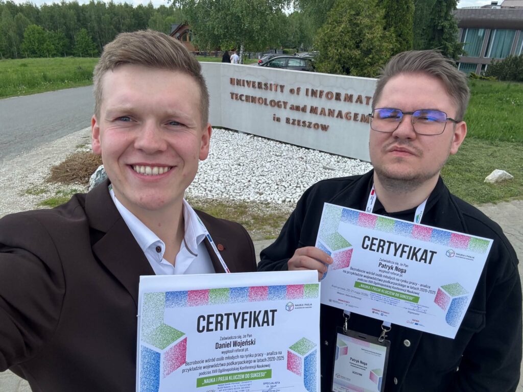 dwóch Studentów stoi z certyfikatami udziału na tle muru z nazwa uczelni