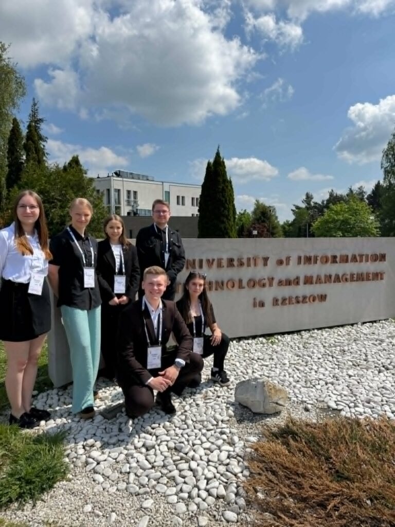 grupa studentów stoi przy dużym murze z nazwa uczelni gościnnej