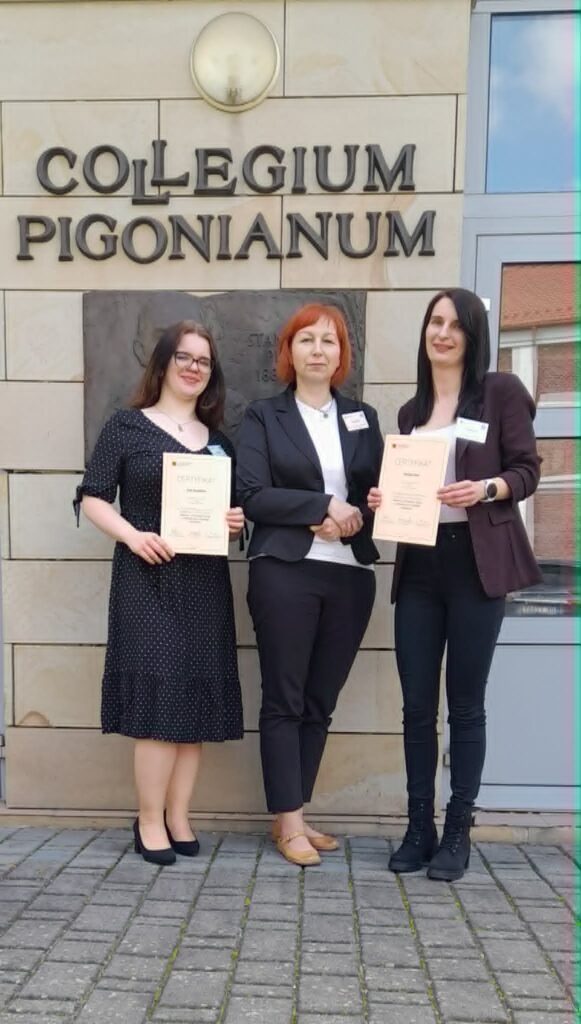 Paulina Szot oraz Julia Kamińska z opiekunem Elżbietą Brągiel stoją z certyfikatami na tle ściany z napisem Collegium Pigonianum