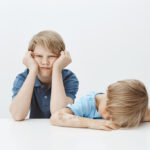 Children feeling bored wanting play instead of doing homework. Portrait of indifferent gloomy brother sitting with sibling, leaning head on hands and squeezing cheeks while boy lying on table. Family concept